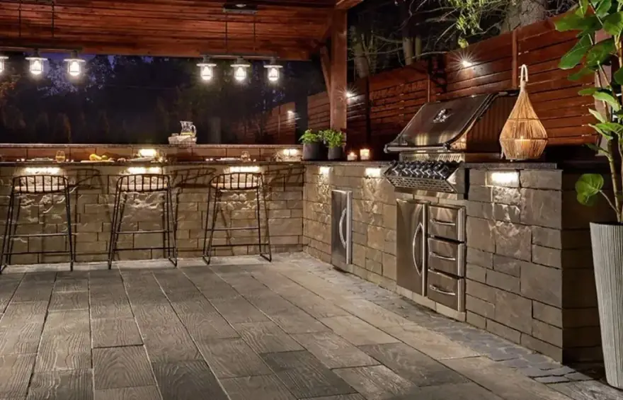 A lit stone outdoor kitchen and bar under a timber pavilion at night, built by Allin Landscaping