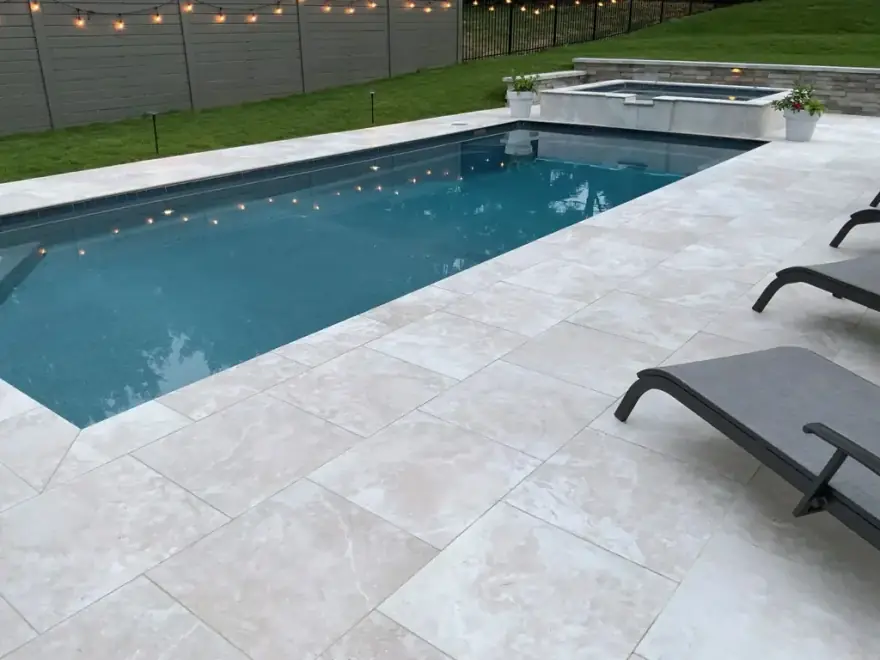 A travertine patio at dusk beside a swimming pool and raised spa strung with festoon lighting